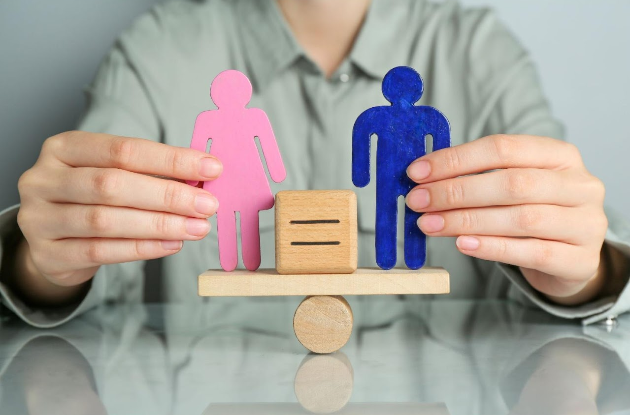 Due mani femminili tengono in equilibrio una piccola bilancia di legno su cui poggiano una sagoma umana rosa e una blu, separate da un blocco di legno con il simbolo dell'uguale, a rappresentare la parità di genere e l'equità salariale.