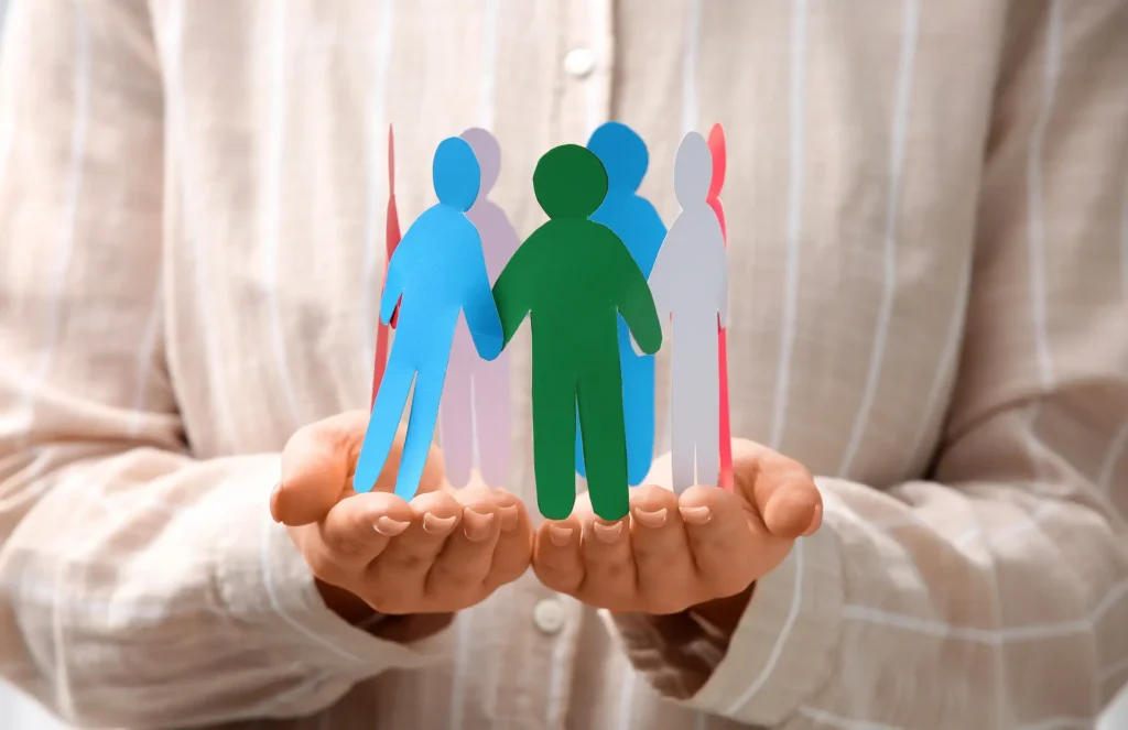 Primo piano di due mani che tengono delicatamente un anello di figure umane di carta colorata collegate tra loro, con una figura centrale verde brillante. Questa immagine simboleggia la comunità, l'inclusione e l'impatto sociale generato dalle organizzazioni, concetto chiave per comprendere la differenza tra il Bilancio Sociale e il Bilancio di Sostenibilità.