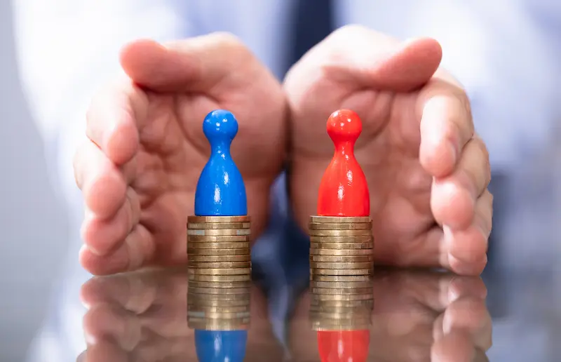 Due pile di monete di altezza diversa con pedine blu e rossa, mani protettive sullo sfondo: rappresentazione del gap retributivo di genere