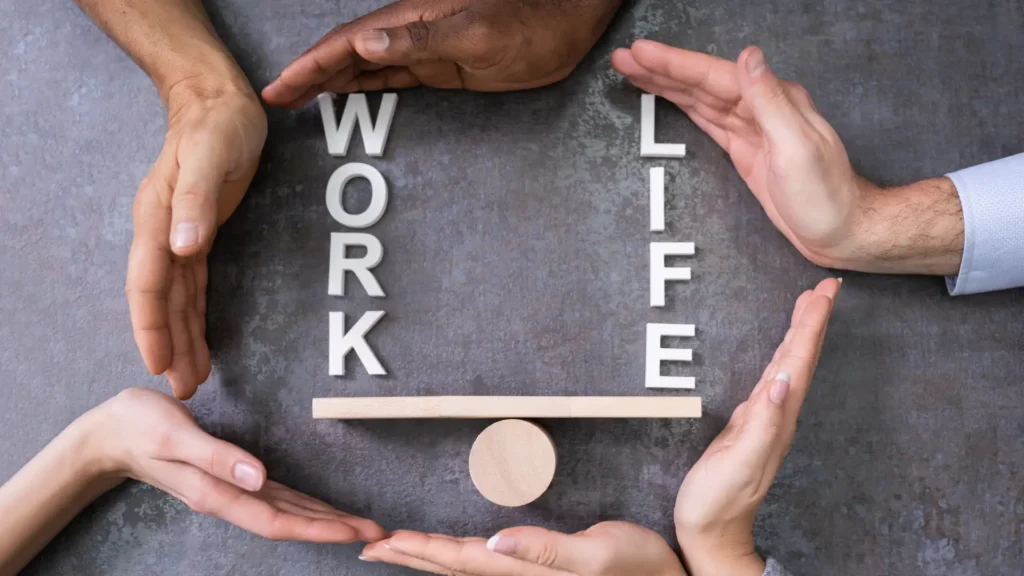 Fotografia dall'alto di quattro mani diverse che proteggono e sostengono una bilancia di legno con le parole 'WORK' e 'LIFE', a simboleggiare l'equilibrio e la conciliazione vita-lavoro supportata dall'organizzazione.