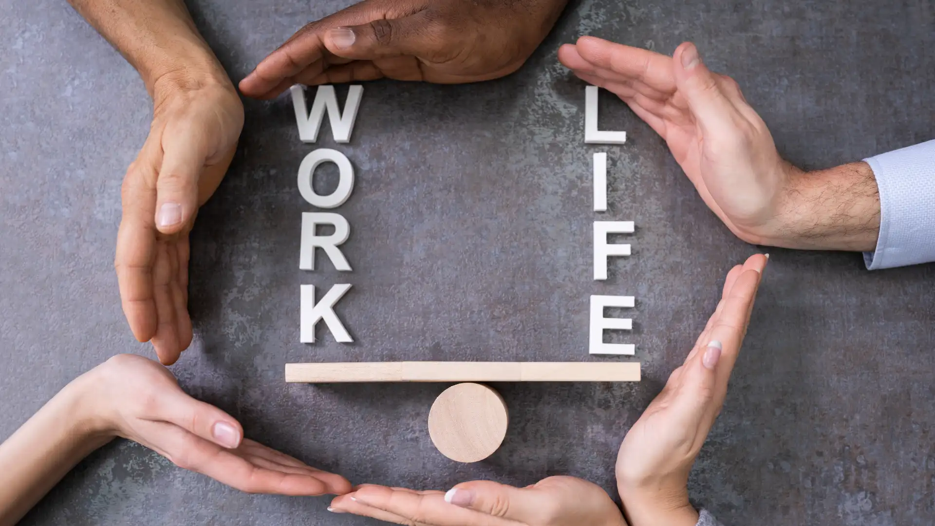 Fotografia dall'alto di quattro mani diverse che proteggono e sostengono una bilancia di legno con le parole 'WORK' e 'LIFE', a simboleggiare l'equilibrio e la conciliazione vita-lavoro supportata dall'organizzazione.