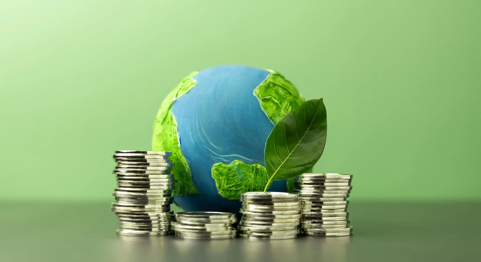 Globo terrestre dipinto in verde e blu accanto a pile di monete con una giovane foglia verde, a rappresentare la crescita economica generata dagli investimenti in sostenibilità ambientale