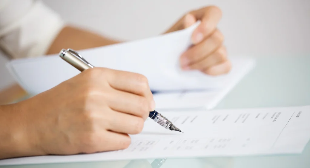 Persona in giacca che firma un documento con una penna stilografica mentre esamina dei fogli, a rappresentare la verifica e la conformità dei processi di due diligence aziendale