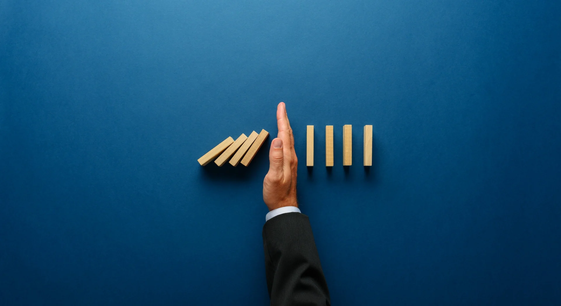Mano di un manager in giacca che blocca una fila di tessere del domino in caduta, a rappresentare la gestione del rischio e la prevenzione degli impatti sull'azienda